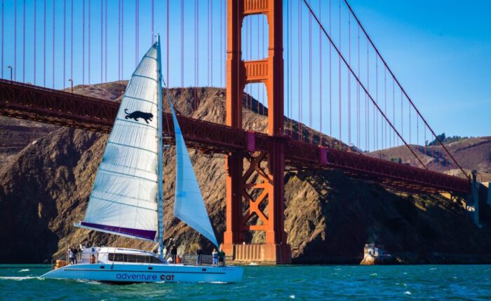 A sailboat named "adventure cat" sails under the golden gate bridge on a sunny day during a San Francisco Bay sailing tour.